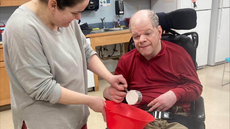 A staff member assists a program participant in a wheelchair with a cooking activity in a community day program kitchen