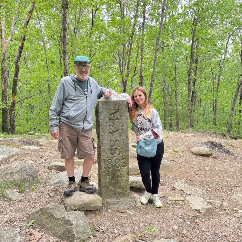 Two people posing next to a Massachusetts state boundary marker during an outdoor community outing
